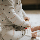 A baby sitting on a white blanket in a nursery, wearing a cream ribbed long-sleeve footless onesie with a small pencil print and holding a white toy.