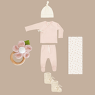 Baby clothing and accessories including a onesie, hat, booties, and a floral ring on a beige background.