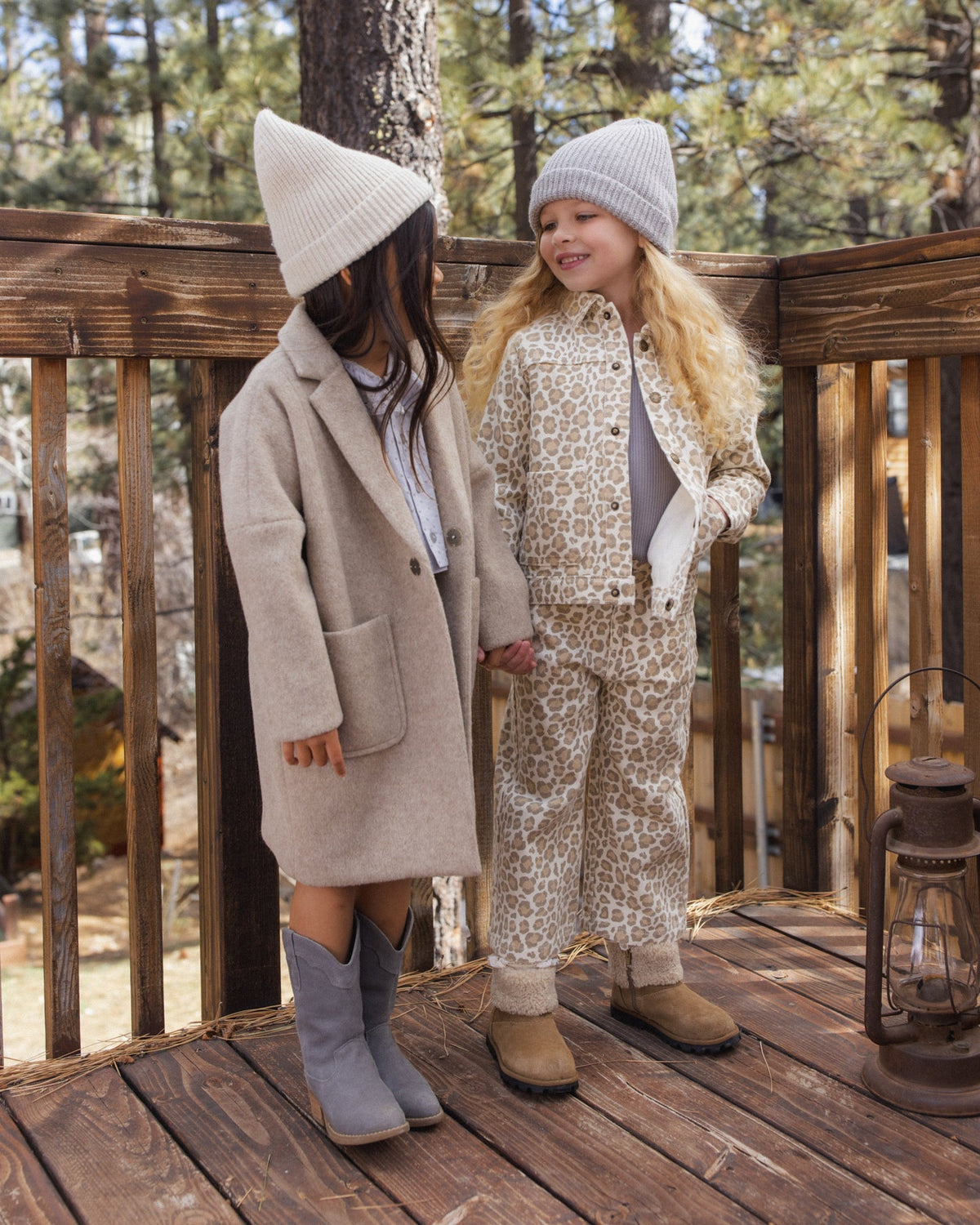 Two young girls standing on a wooden deck outdoors, wearing coordinated fall fashion. One girl wears a beige coat with a matching beanie and gray boots, while the other wears a beige-and-white animal-print jacket and matching pants with a light knit beanie and tan boots.