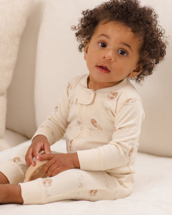 A baby with curly dark hair sits on a white sofa wearing a cream-colored long-sleeve footless sleeper with a zipper and a small brown animal print.