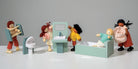 A lifestyle product photo featuring the Wooden Bathroom Furniture set by Mentari alongside several dollhouse dolls interacting with the pieces against a white background. One doll carries a wooden towel stand, another stands next to the toilet, a third washes their hands at the sink unit with a mirror, a fourth relaxes in the mint-green bathtub, and a fifth carries a pink fabric laundry basket.
