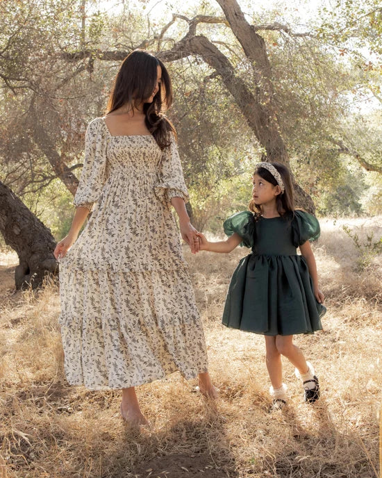 A woman and a young girl holding hands walk through a dry grassy outdoor area with trees. The woman wears a cream floral tiered maxi dress, and the girl wears a dark green puff-sleeve dress.