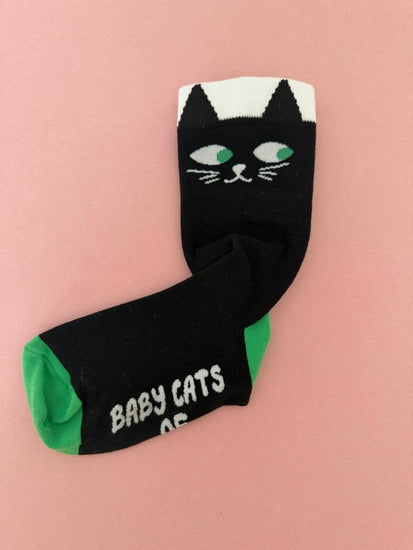 A pair of black novelty socks featuring a black cat face design on the cuff and the words 'BABY CATS' in white on the toe, with green heels and a pink background.