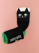 A pair of black novelty socks featuring a black cat face design on the cuff and the words 'BABY CATS' in white on the toe, with green heels and a pink background.