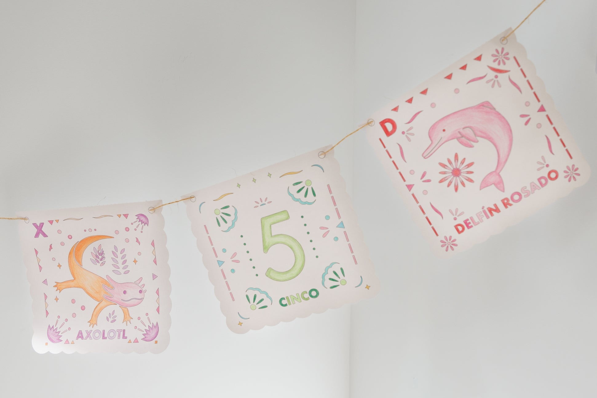 Three decorative hanging pennants strung together against a plain light wall. The center pennant shows the number 5 in green with the word Cinco, and the side pennants have pastel illustrations with text including AXOLOTL and DELIN ROSSO.