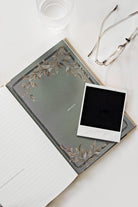 Green notebook with floral design on cover, white photo frame, and glasses on a light surface