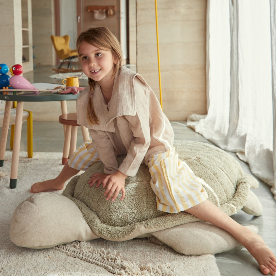 Child sitting on a large cushion in a bright, well-lit room with toys and furniture.