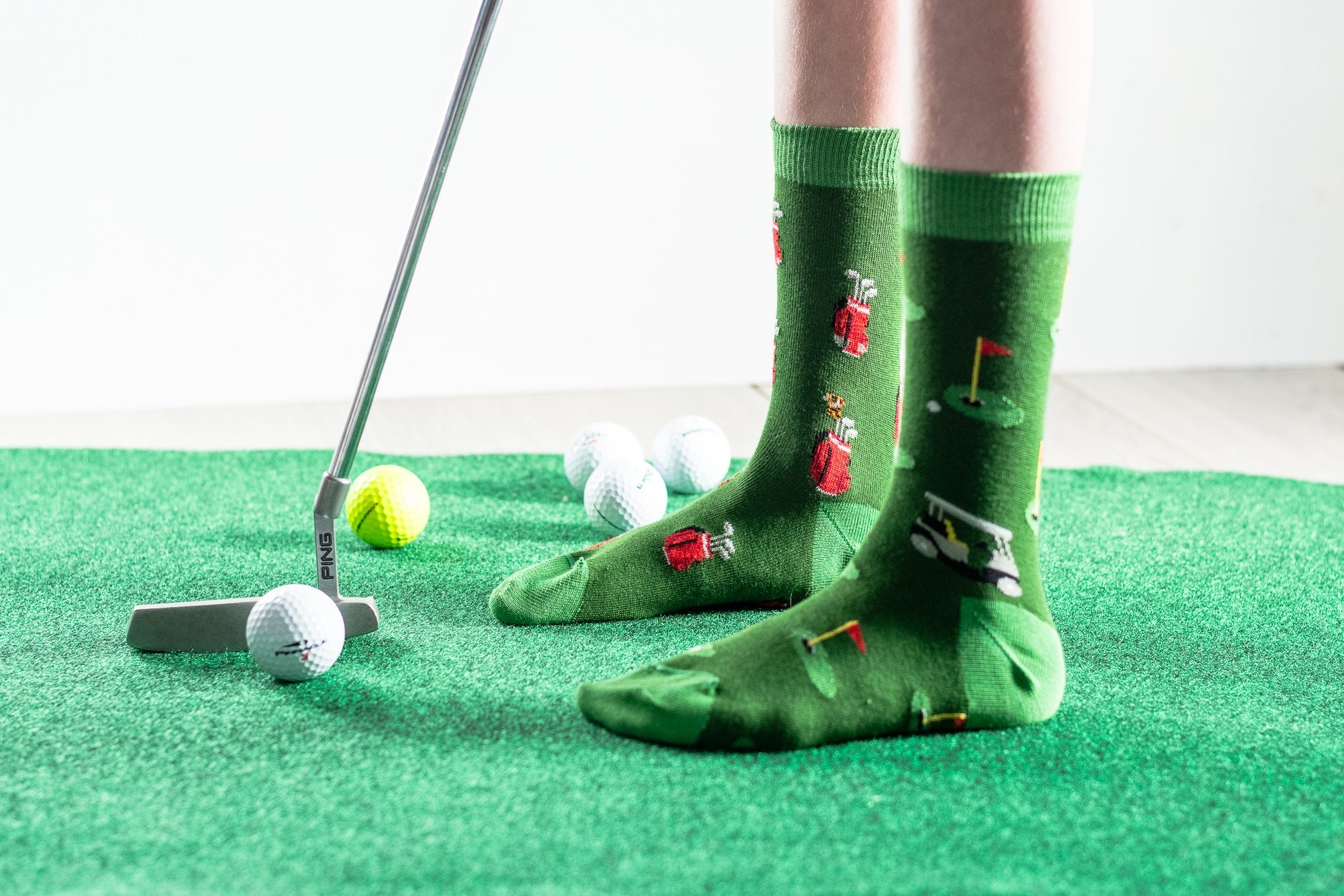 A person wearing green novelty golf socks with illustrated golfers and clubs, standing on a green turf mat with golf balls and a putter nearby.