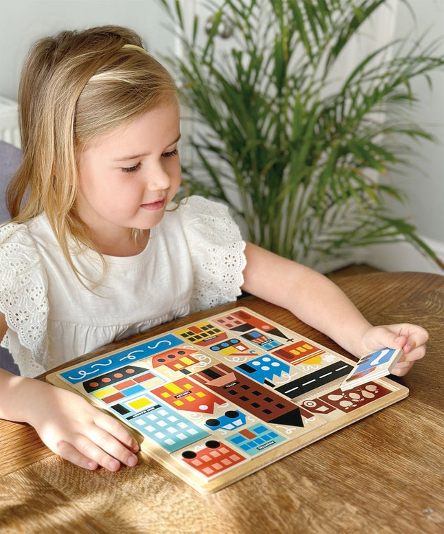 A young girl with blonde hair tied back sits at a wooden table, smiling as she plays with the Mentari Wooden Town Puzzle, holding a puzzle piece in her hand while assembling the colorful town scene.