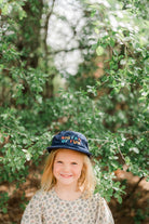 Child wearing a cap with text, standing in a forest setting