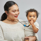 A baby wearing a cream ribbed short-sleeve shirt holds a black silicone teether with a blue and tan accent against their cheek, with a mother's face partially visible on the left.
