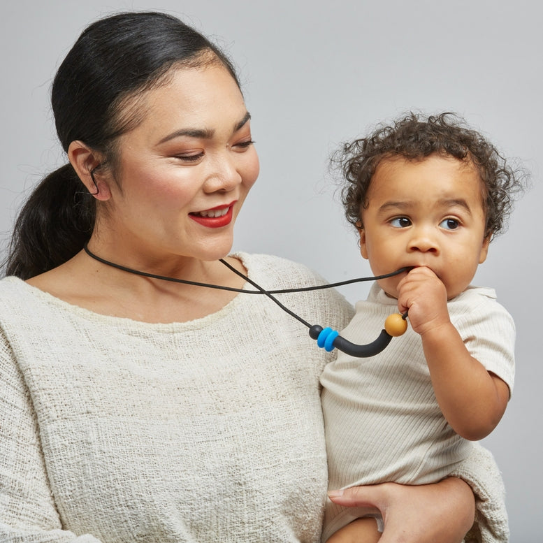 A baby wearing a cream ribbed short-sleeve shirt holds a black silicone teether with a blue and tan accent against their cheek, with a mother's face partially visible on the left.