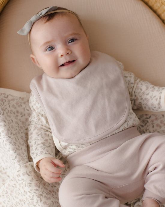 A smiling baby wearing a light beige bib over a matching beige and white patterned long-sleeve top and beige pants, lying on a soft beige blanket.