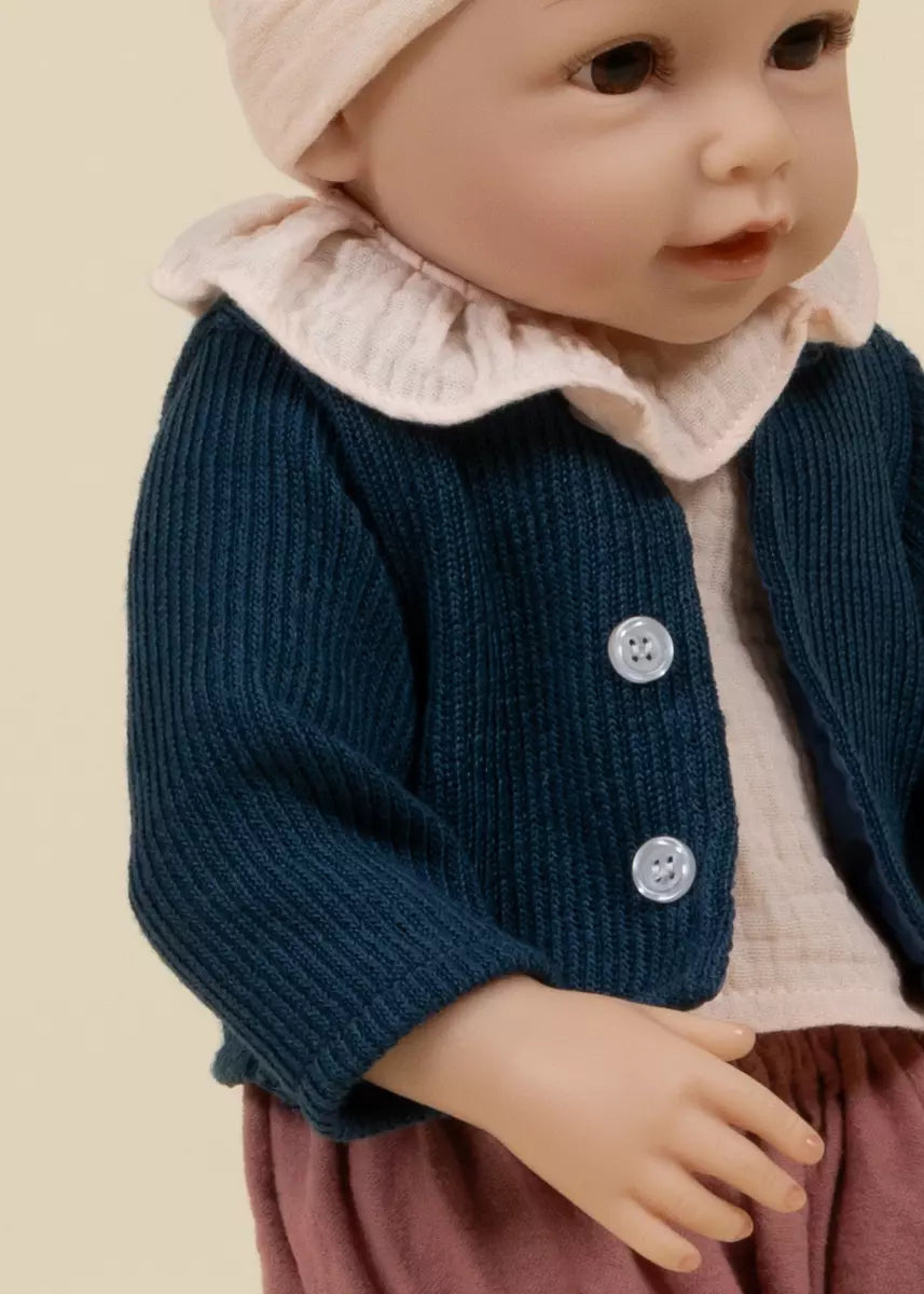 Baby doll wearing a blue cardigan, pink shirt, and beige hat on a beige background