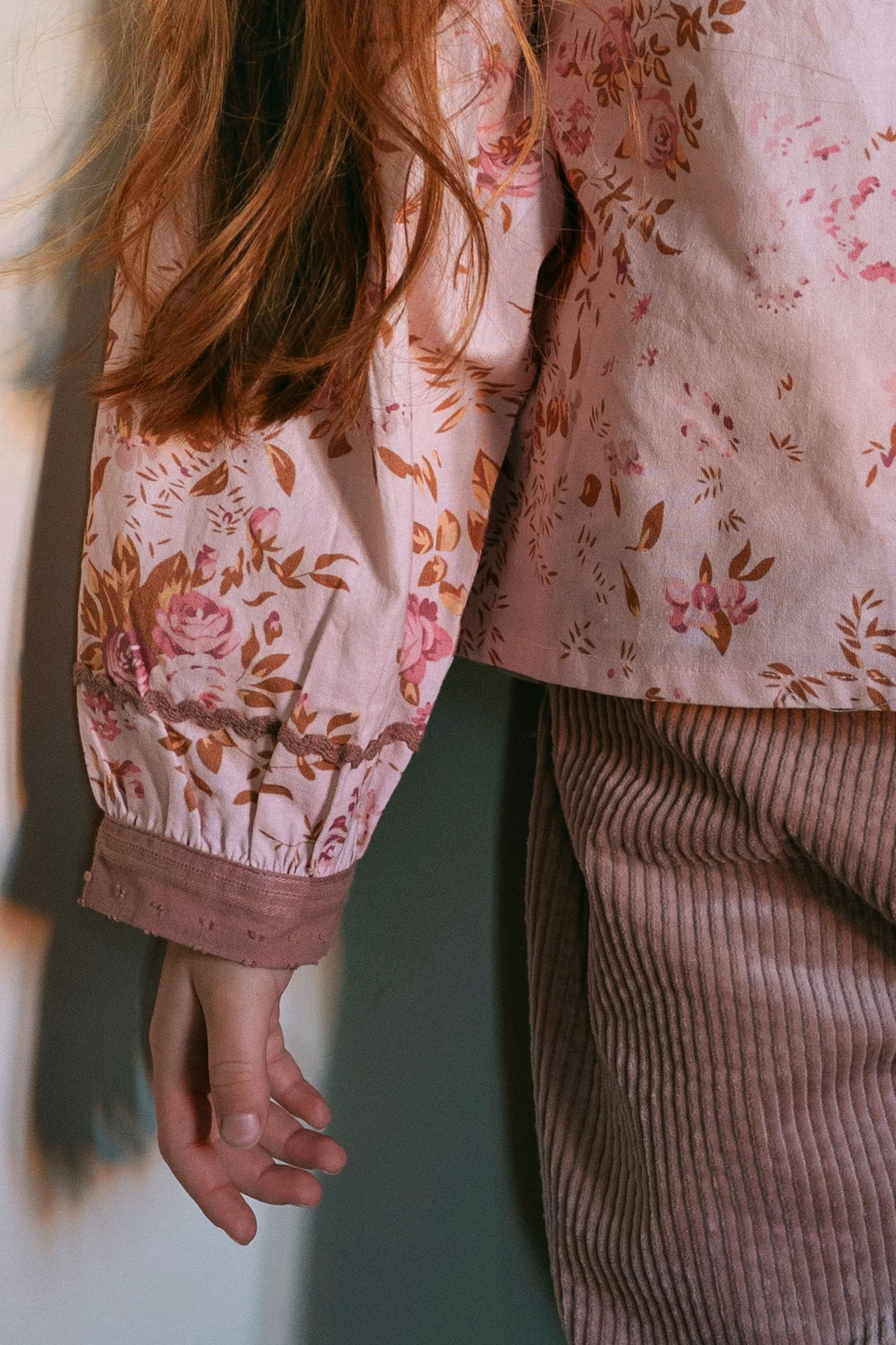 A close-up lifestyle shot of a person wearing a light pink floral long-sleeve top with brown lace trim and a dusty rose corduroy skirt, standing against a neutral wall.