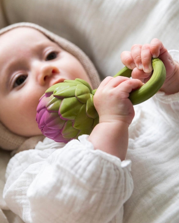Baby lying down in a white ruffled outfit, holding the pink-and-green Oli & Carol artichoke rattle toy and exploring it with their hands.