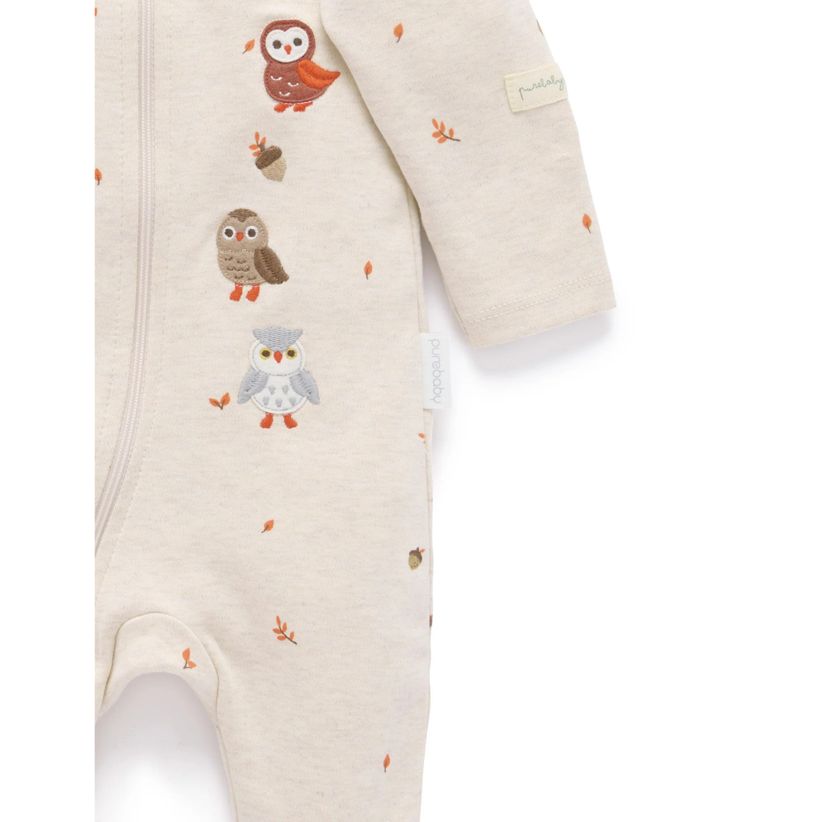 A close-up of a baby footed one-piece romper in a warm cream heather fabric, decorated with a vertical pattern of colorful owls and small orange acorns and leaves. The brand name 'prouvenc' is visible on a small fabric tag on the sleeve.