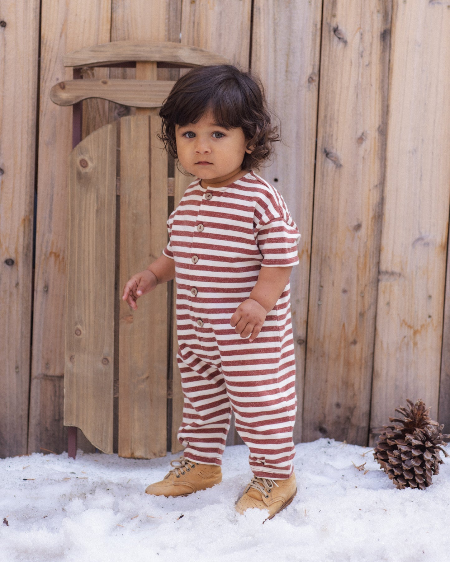 A toddler stands outdoors in the snow in front of a wooden fence, wearing a red and white horizontally striped one-piece jumpsuit with snap buttons and tan boots.