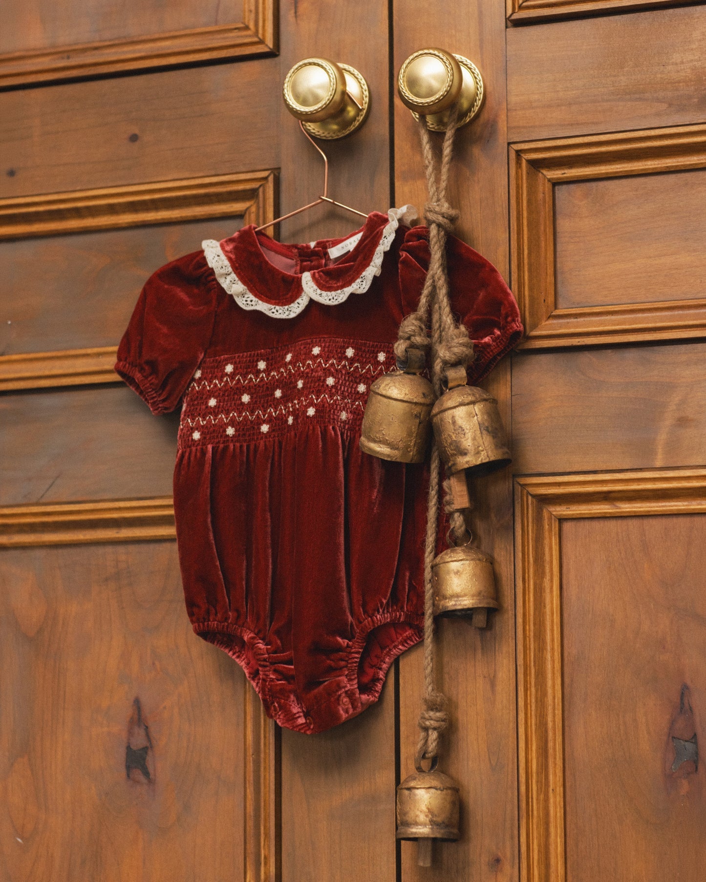 A baby's short-sleeve velvet romper in deep burgundy red, featuring a white embroidered Peter Pan collar and white dot embroidery across the chest, hanging on a wooden door with brass bell charms.