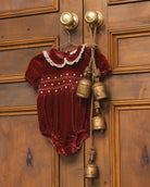 A baby's short-sleeve velvet romper in deep burgundy red, featuring a white embroidered Peter Pan collar and white dot embroidery across the chest, hanging on a wooden door with brass bell charms.