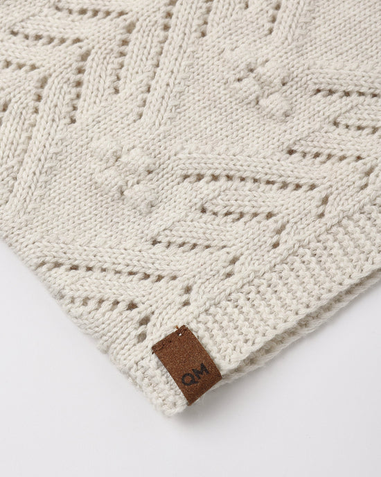 Close-up of a cream-colored knit blanket with a geometric cable-knit pattern and a small brown leather brand tag reading 'QM'.