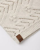 Close-up of a cream-colored knit blanket with a geometric cable-knit pattern and a small brown leather brand tag reading 'QM'.