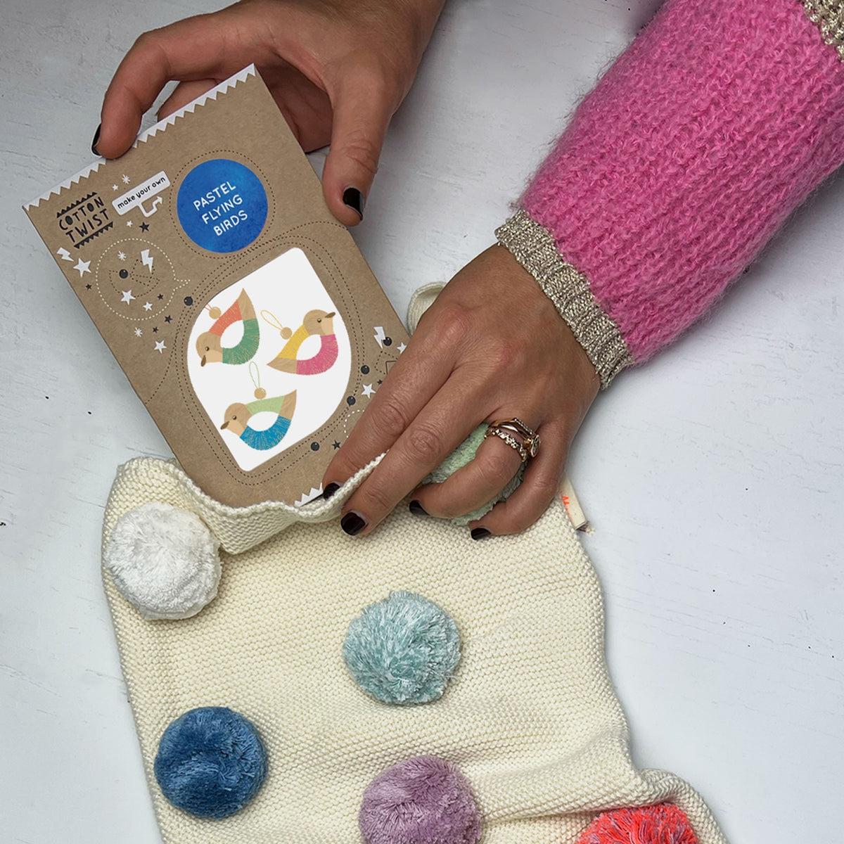 Person holding a package of pom-pom making kit with colorful pom-poms on a white surface