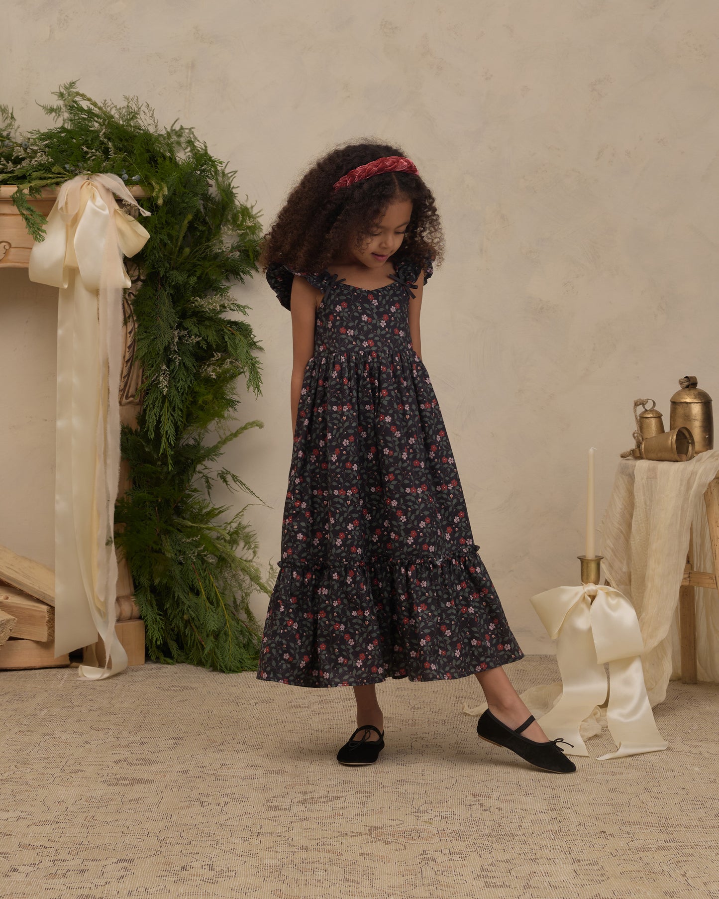 A young girl with curly hair wearing a dark floral midi dress, black flats, and a red headband, standing in a styled studio setting with a textured beige wall, greenery, and cream ribbons.