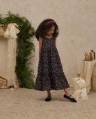A young girl with curly hair wearing a dark floral midi dress, black flats, and a red headband, standing in a styled studio setting with a textured beige wall, greenery, and cream ribbons.