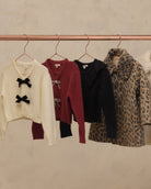 Four women's knit outerwear pieces hanging on a copper-colored clothing rack against a textured beige wall. From left to right: a cream cropped sweater with black bow details, a burgundy cropped sweater with white tie details, a black ruffled cardigan, and a leopard-print coat.