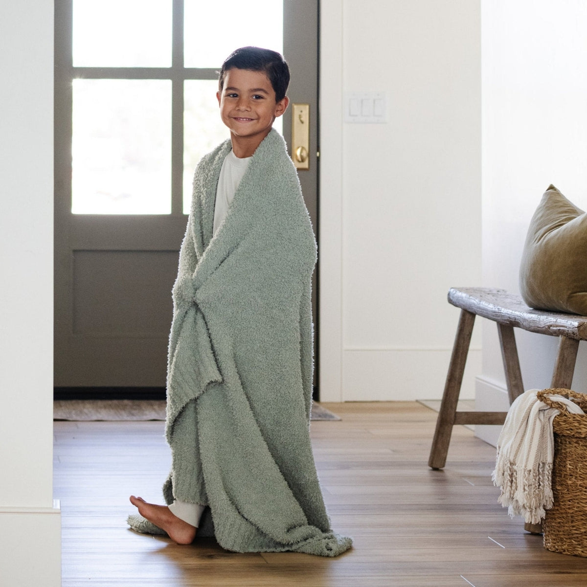 Child wrapped in a green blanket standing in a room with a wooden floor and a door.