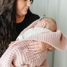 A close-up lifestyle shot of a woman holding a sleeping newborn baby wrapped in a dusty pink textured knit blanket and wearing a white knit hat. The woman is wearing a black top and a gold ring.