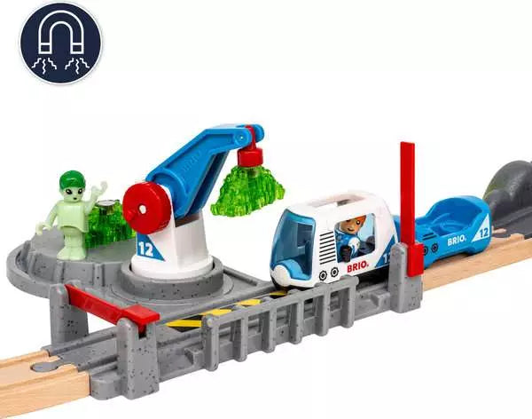 A Space Deluxe Set by Brio astronaut figure standing near a grey railway crossing gate, a white and blue space train, a blue cargo crane lifting a green crystal, and a glow-in-the-dark alien standing next to a grey rocky terrain piece.