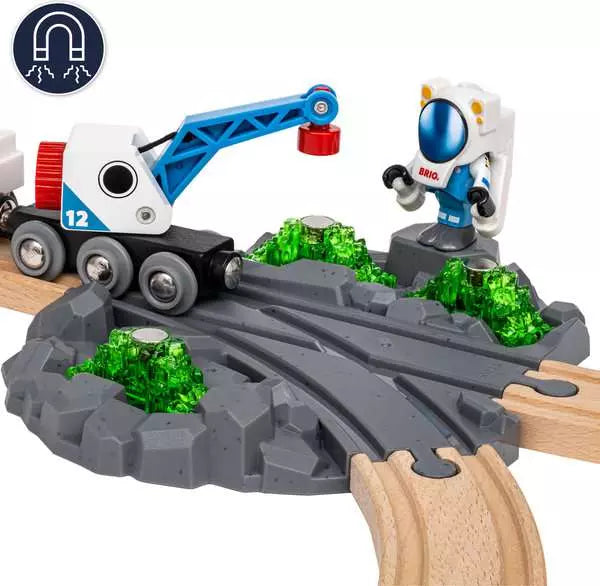 A Space Deluxe Set by Brio astronaut figure standing near a grey railway crossing gate, a white and blue space train with a blue cargo crane, a green glowing crystal on a rocky terrain piece, and a circular magnet icon in the top-left corner.