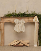 An ivory baby dress with puffed sleeves and a large bow is hanging from the center of an ornate wooden mantel. The mantel is decorated with green foliage, three tall candles, and a long cream ribbon, with stacked firewood below against a textured beige wall.