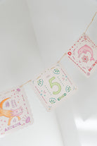 A close-up of a decorative banner with three triangular flags hanging on a string against a white wall. The center flag says “5” and “Díaá” in green and pink lettering, decorated with small green and pink motifs.