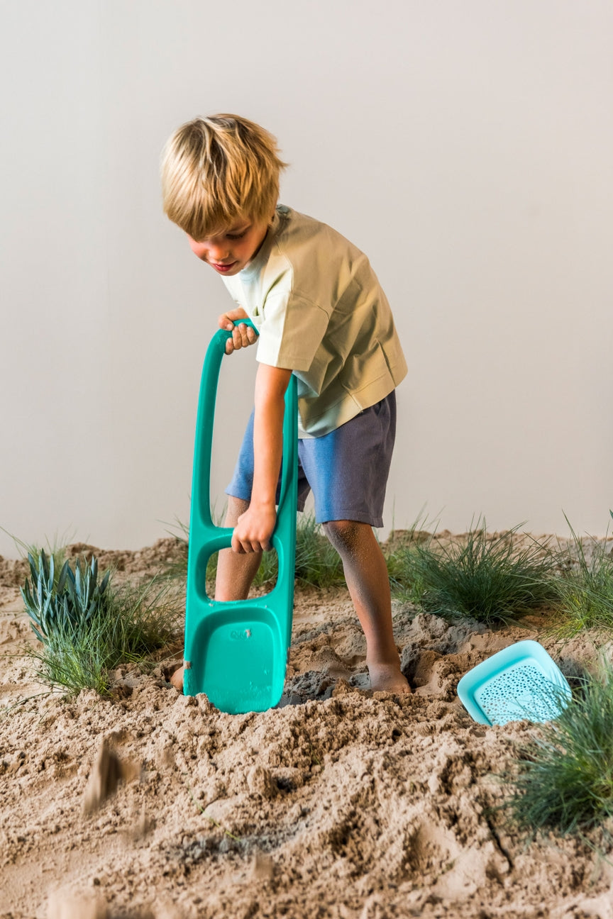 Child playing in the sand with a green toy shovel and blue sandcastle mold.