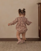 A toddler girl seen from behind wearing a long-sleeve floral romper and pink ballet shoes, standing on a patterned rug against a beige wall.