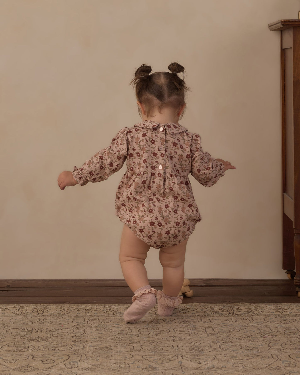 A toddler girl seen from behind wearing a long-sleeve floral romper and pink ballet shoes, standing on a patterned rug against a beige wall.