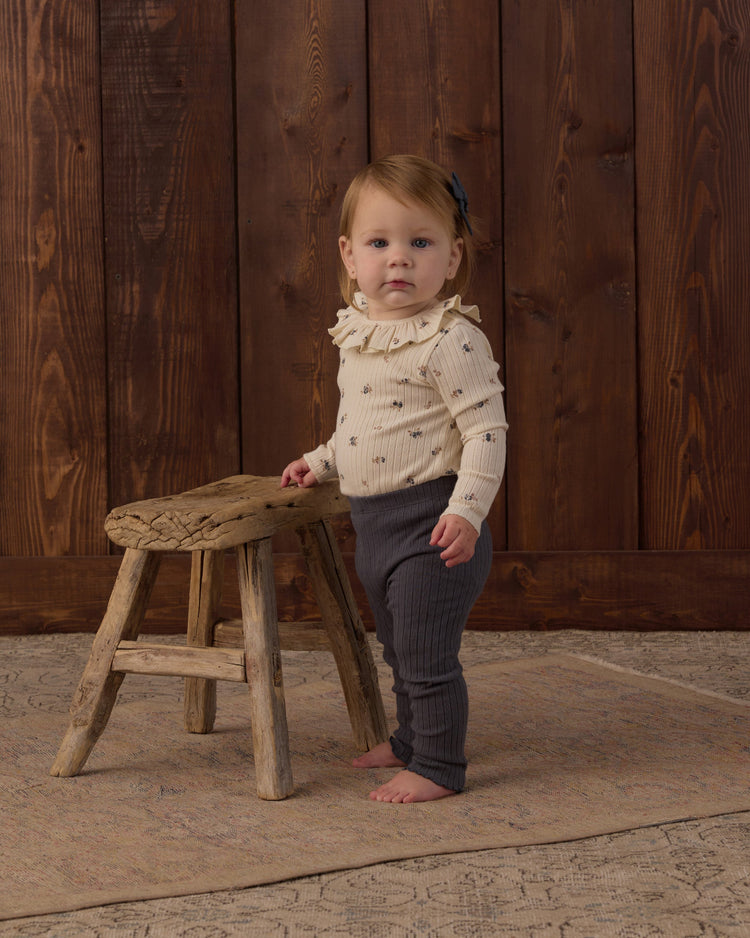 A toddler wearing Rylee + Cru Ruffle Collar Bodysuit in Natural Ditsy paired with Midnight ribbed leggings and a matching hair bow.