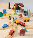 A top-down, close-up view of a wooden toy train set on a light-colored surface, featuring a curved track with a blue train engine and yellow cargo cars. Colorful wooden town buildings, including a red house, blue structures, and abstract trees, surround the tracks alongside a yellow traffic light, a red toy car, and a yellow toy truck.