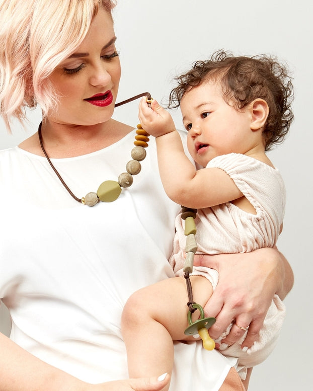 Woman holding a baby and wearing a necklace with large beads on a plain background.