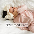 Baby wearing a pink outfit and white headband with a 'Trimmed Knot' label.