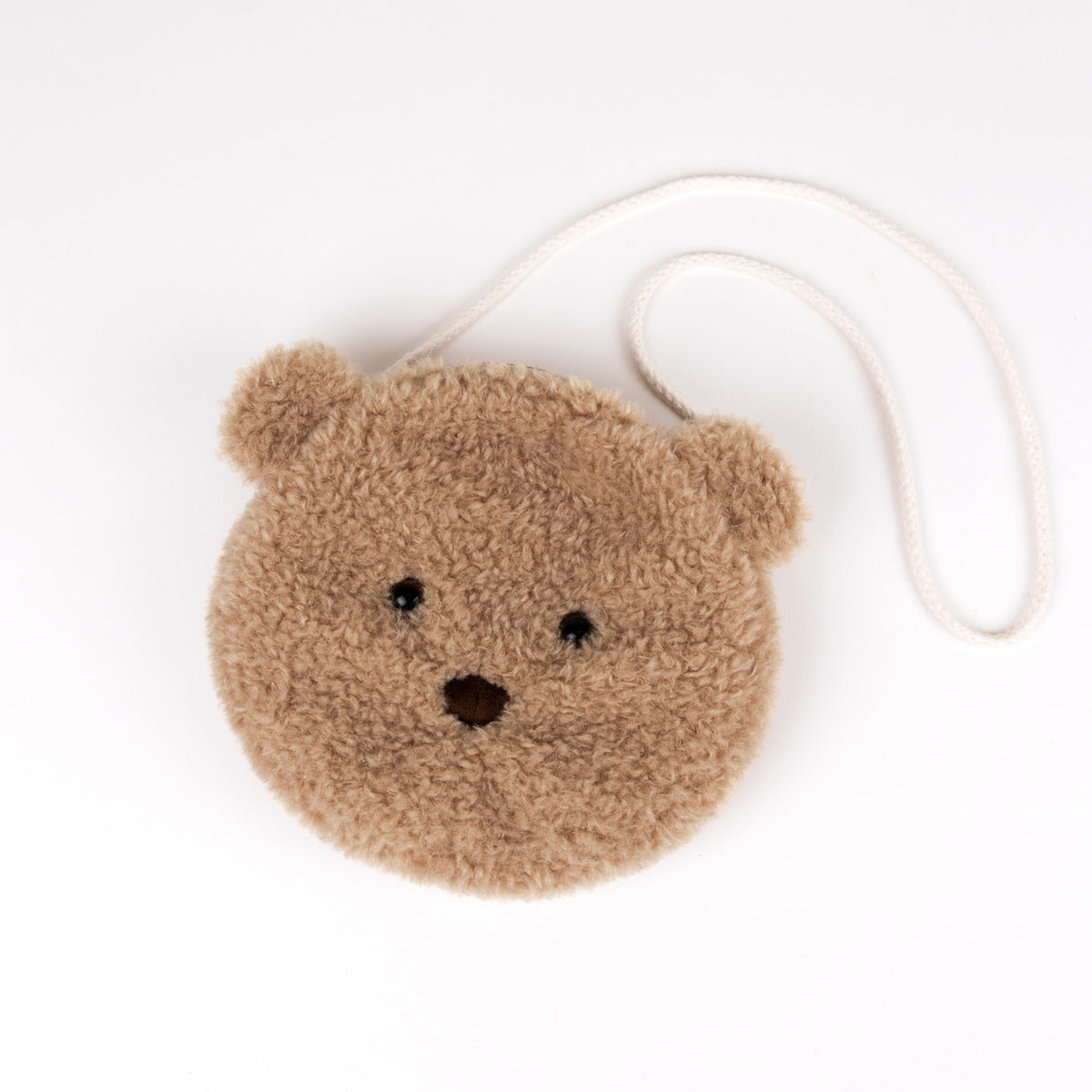 A round, plush teddy bear face crossbody bag in a warm tan-brown color, featuring embroidered black eyes and nose, with a small ear on each side. It has a white braided shoulder strap and is photographed on a clean white background.