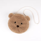 A round, plush teddy bear face crossbody bag in a warm tan-brown color, featuring embroidered black eyes and nose, with a small ear on each side. It has a white braided shoulder strap and is photographed on a clean white background.