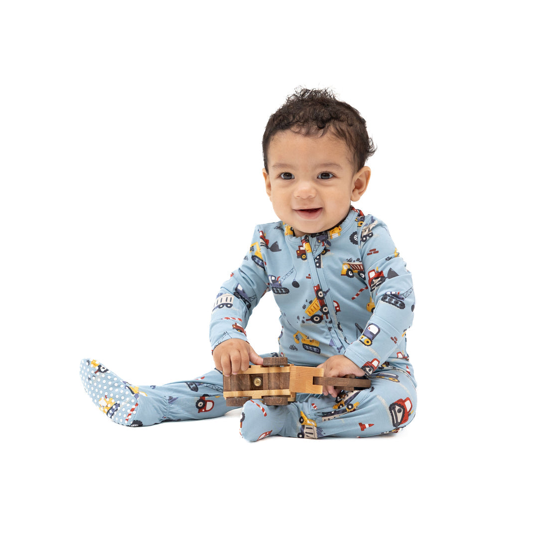 A baby sitting on a white background wearing a light blue one-piece pajama with a construction vehicle print and holding a small wooden toy truck.