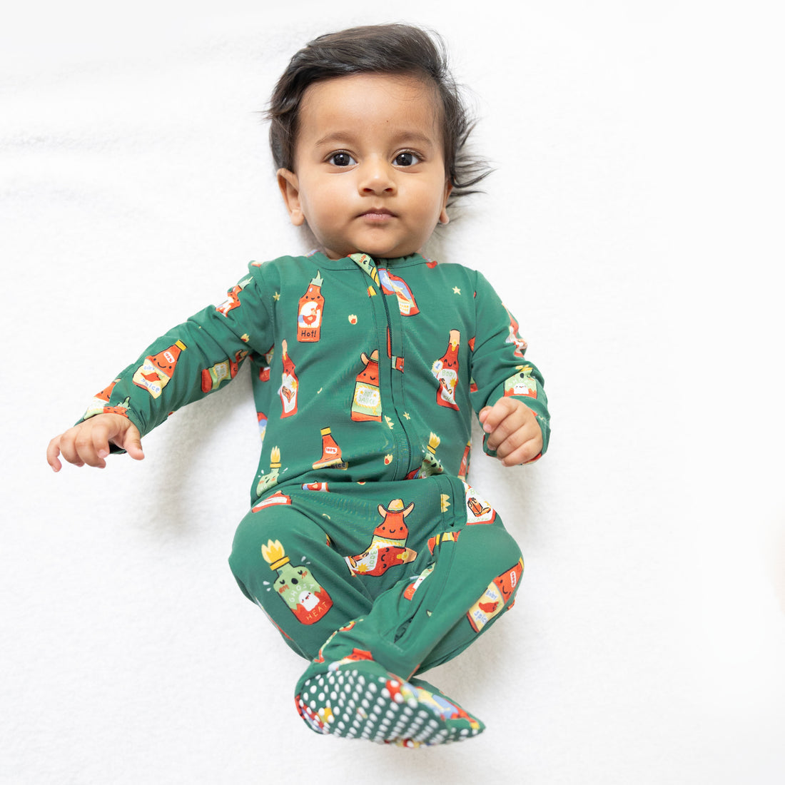 A baby wearing a green long-sleeve footed sleeper with a festive cartoon print, lying on a white textured blanket.