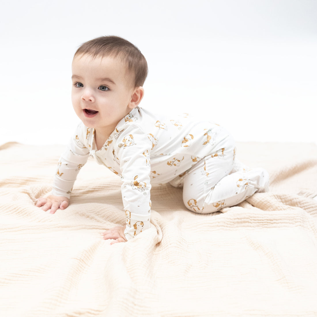 Baby in a white onesie with gold patterns sitting on a beige blanket.