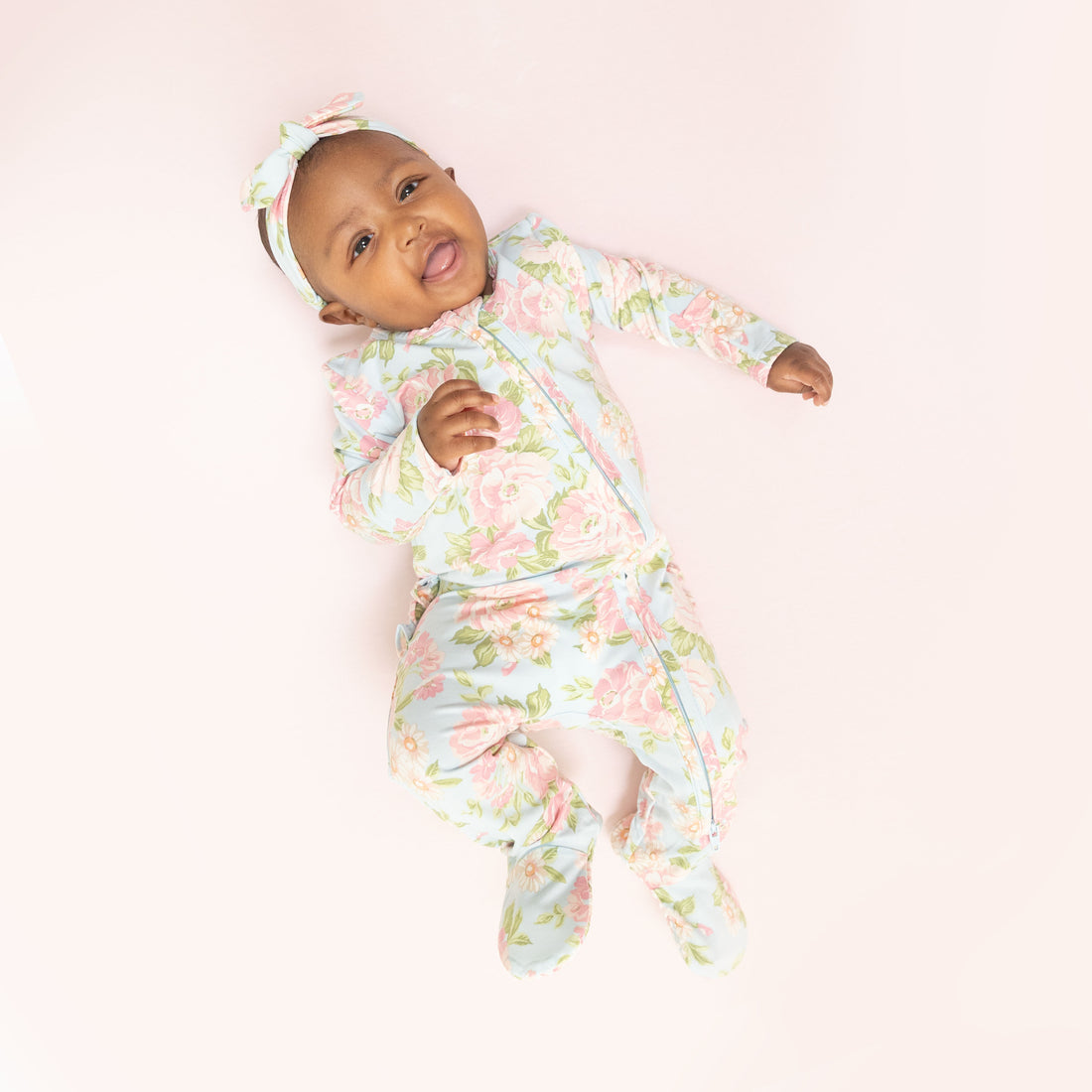 A baby lying on their back wearing a light blue footed sleeper with a pink and green floral pattern, matching floral headband, and smiling with their tongue sticking out, on a solid pale pink background.