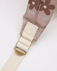 Close-up of a shoulder strap with a dusty pink fabric body featuring a brown floral pattern, an off-white woven strap, and a gold-toned metal buckle.